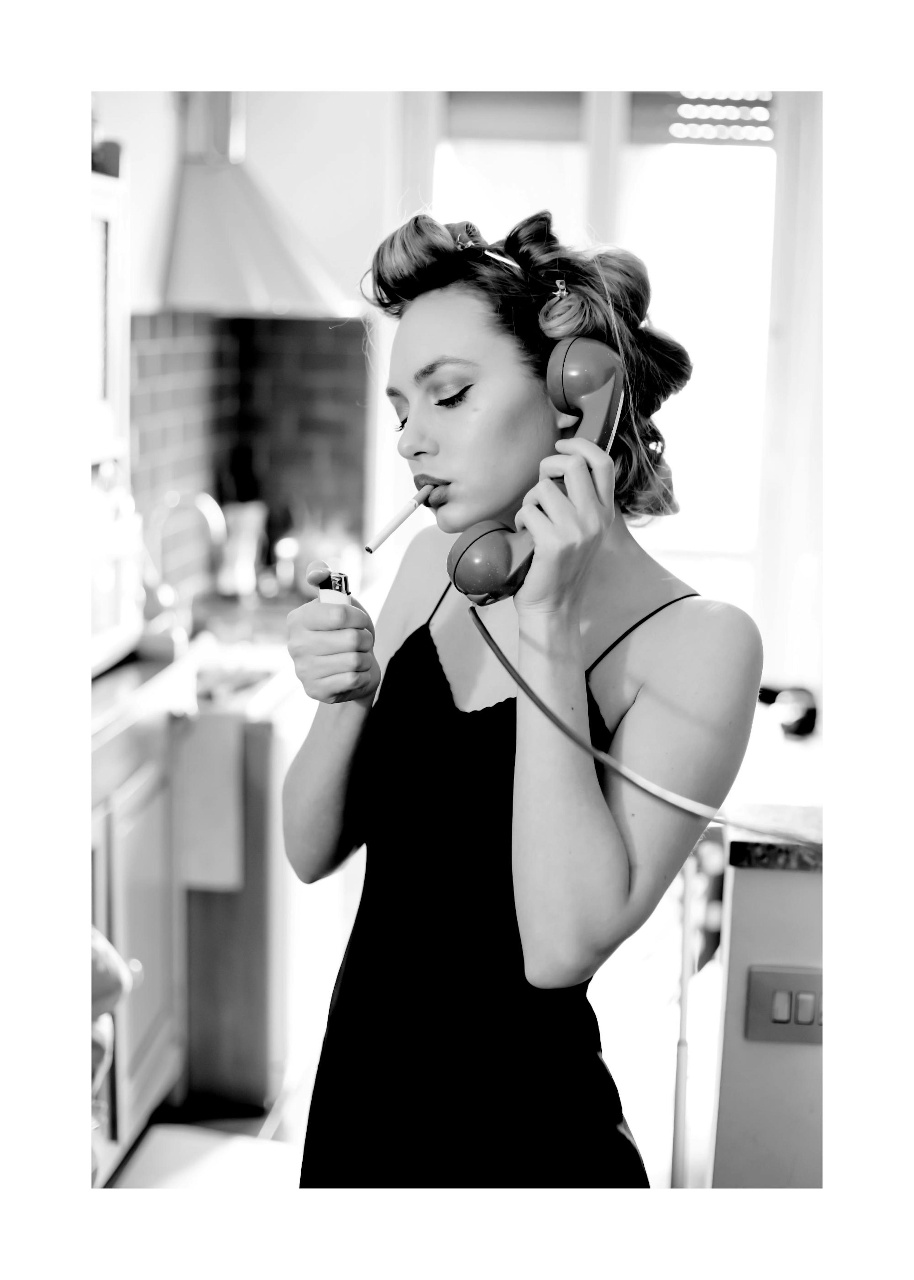 Čiernobiely plagát ženy s natáčkami, ktorá telefonuje, drží cigaretu a zapaľovač v retro kuchyni.
