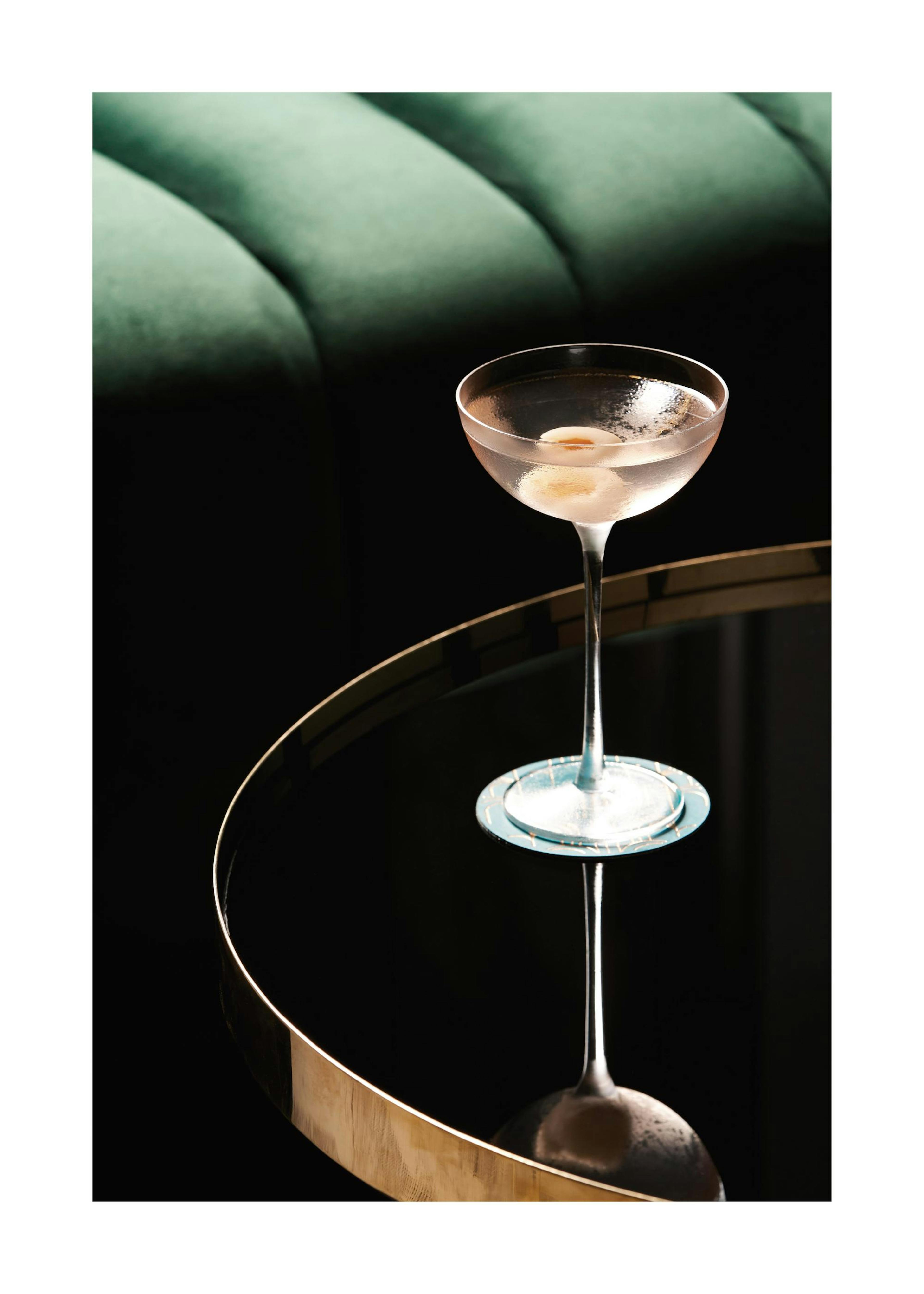 A print of a pink cocktail on a gold-rimmed table, displayed above a white cabinet with a glass vase and open book.