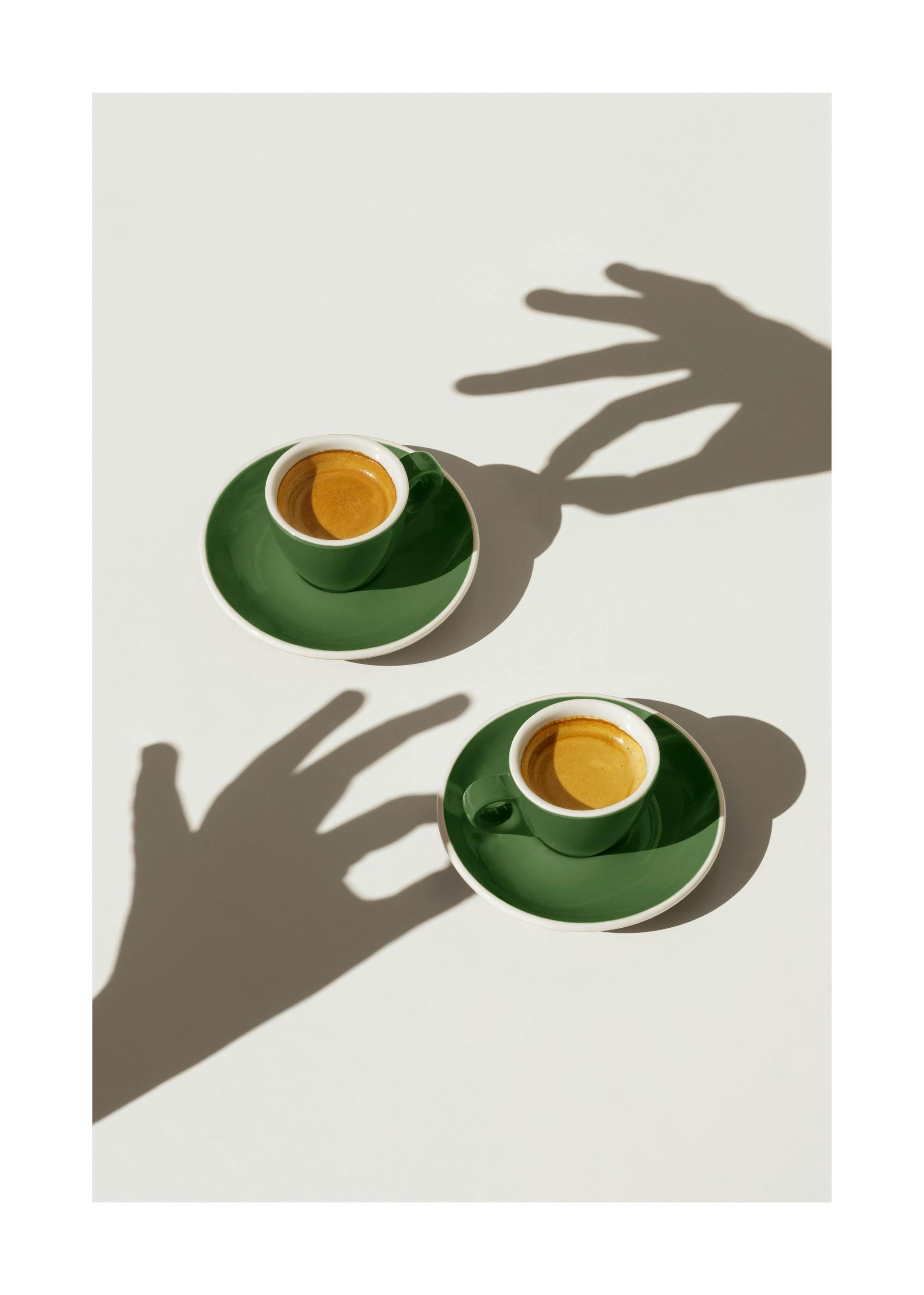 Zwei grüne Kaffeetassen mit Untertassen auf weißem Hintergrund und zwei Schattenspielen von Händen, die nach den Tassen greifen.